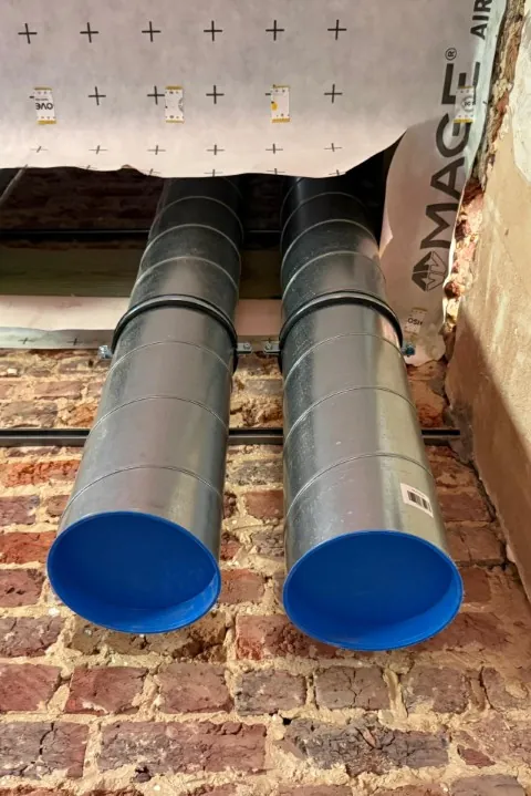 Twee gegalvaniseerde ventilatiekanalen met blauwe afdekkappen tegen een bakstenen muur
